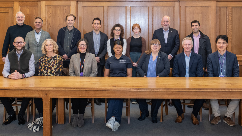 A group of people some sitting in front of a wooden table, some standing behind, in a wood panelled room